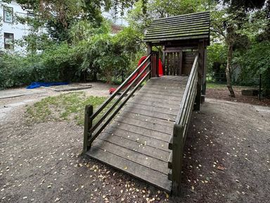 Hölzernes Spielhaus mit Rampe und roter Rutsche, daneben Sandkasten und Rasen unter hohen Bäumen